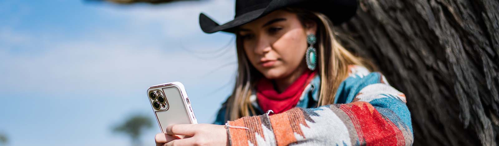 A woman sitting against a tree, looking at her cellphone.