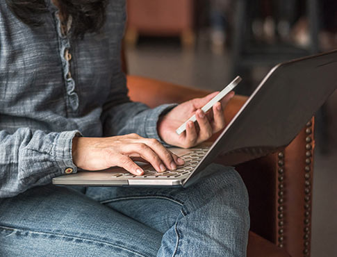 An image of a woman looking at her phone and typing on a laptop.
